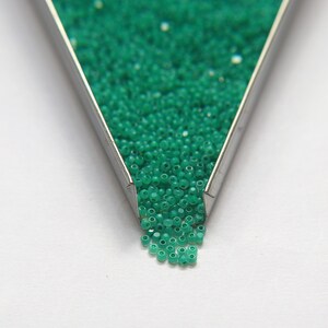 Op de afbeelding: Een metalen bakje gevuld met kleine, felgroene glazen kralen. De kralen vallen uit het bakje op een wit oppervlak.