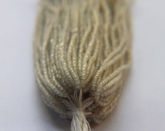 May include: Close-up of a bundle of small, light-colored beads. The beads are strung together, forming multiple strands that are tied together at the bottom with a white string. The beads have a slightly iridescent quality.