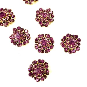 Könnte beinhalten: Sieben runde Broschen oder Knöpfe im Vintage-Stil. Jedes hat einen zentralen, großen, runden, lila Strassstein, der von kleineren lila Strasssteinen umgeben ist. Die Fassungen sind goldfarben. Die Broschen sind auf weißem Hintergrund angeordnet.