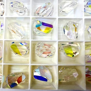 May include: A selection of clear, faceted crystal beads displayed in a white compartmented tray. The crystals shimmer with a spectrum of colours, including yellow, blue, and purple. Each bead is multi-faceted, producing a sparkling effect. These beads are likely for jewellery making or crafting.