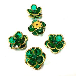 Può includere: Cinque abbellimenti a forma di fiore con pietre sfaccettate verde smeraldo e montature in metallo dorato. I disegni floreali sono disposti su uno sfondo bianco, mostrando i loro dettagli intricati e il colore vibrante.