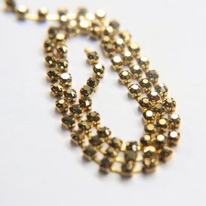 Puede incluir: Una cadena dorada hecha de pequeños diamantes de imitación facetados. La cadena está en bucle y se encuentra sobre una superficie blanca.