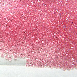 May include: A close-up view of a pile of small, faceted, pink crystals. The crystals are scattered across a white surface, with varying degrees of light reflection. The crystals appear to be uniform in size and shape, suggesting they are manufactured.