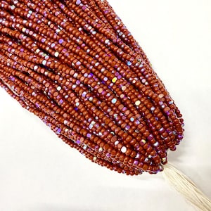 May include: Close-up of a bundle of small, reddish-brown beads with iridescent highlights. The beads are strung together, forming a dense, textured mass. A white tassel is visible at the base of the bundle. The beads appear to be made of a shiny material.