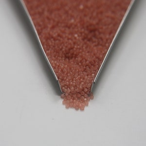 Peut inclure: Gros plan d'une cuill&egrave;re en m&eacute;tal remplie de petites perles de verre rose clair. Les perles se r&eacute;pandent de la cuill&egrave;re sur une surface blanche.