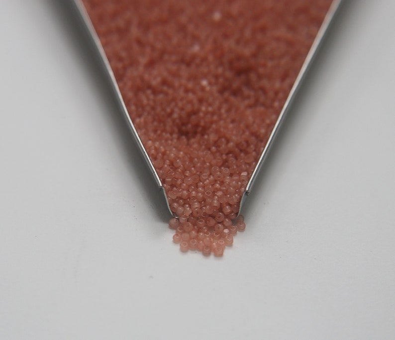 Peut inclure: Gros plan d'une cuill&egrave;re en m&eacute;tal remplie de petites perles de verre rose clair. Les perles se r&eacute;pandent de la cuill&egrave;re sur une surface blanche.