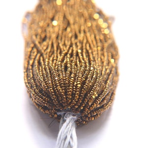 Puede incluir: Primer plano de un paquete de cuentas de semillas doradas. Las cuentas son peque&ntilde;as y redondas, y tienen un brillo met&aacute;lico.