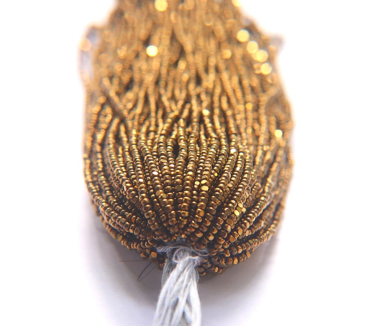 Puede incluir: Primer plano de un paquete de cuentas de semillas doradas. Las cuentas son peque&ntilde;as y redondas, y tienen un brillo met&aacute;lico.