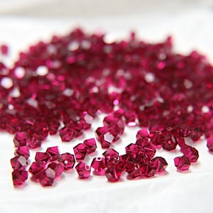 May include: A collection of deep magenta beads with multiple facets, scattered on a white surface. The beads are small, with many flat surfaces that reflect light, creating a sparkling effect. The beads are scattered.