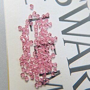 May include: A scattering of small, faceted, light pink beads. The beads are scattered on a white surface with large gray text. The beads have a sparkling, translucent appearance, suggesting they are made of glass or crystal.