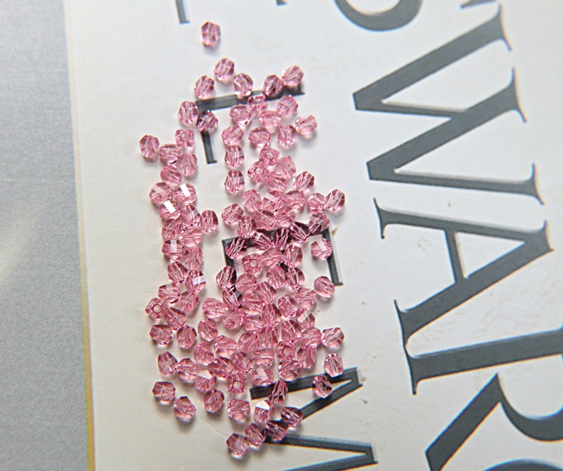 May include: A scattering of small, faceted, light pink beads. The beads are scattered on a white surface with large gray text. The beads have a sparkling, translucent appearance, suggesting they are made of glass or crystal.