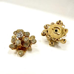 Può includere: Un paio di orecchini a clip dorati vintage. Ogni orecchino presenta un gruppo di strass rotondi e trasparenti incastonati in un motivo floreale. Gli orecchini hanno un aspetto dettagliato e strutturato.