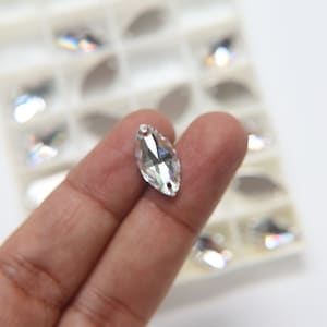 May include: A clear, marquise-shaped crystal rhinestone with facets, held in a hand. The rhinestone has two small holes at each end. Other rhinestones are visible in the background, arranged in a white plastic organizer.