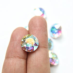 Op de afbeelding: Een close-up van regenboogkleurige ronde strasssteentjes, met één strasssteentje vastgehouden tussen twee vingers. De strasssteentjes hebben een regenboogeffect.