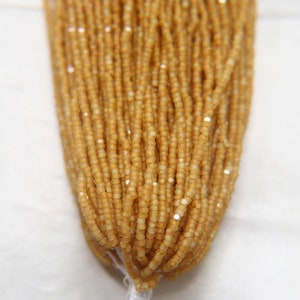 May include: A strand of small, golden-brown seed beads with a subtle shimmer. The beads are uniform in size and shape, creating a smooth, continuous texture.