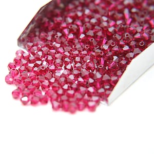 Puede incluir: Primer plano de una gran cantidad de pequeñas cuentas de vidrio facetadas de color rojo rubí. Las cuentas están en una cuchara de metal, derramándose sobre una superficie blanca.