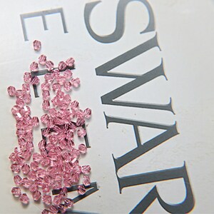 May include: A collection of small, faceted, light pink crystal beads scattered on a white surface. The beads are clustered together, reflecting light and creating a sparkling effect. The word "SWAROVSKI" is partially visible in the background.
