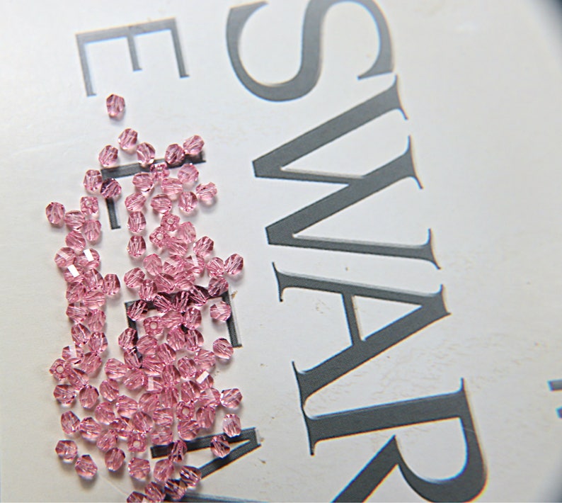 May include: A collection of small, faceted, light pink crystal beads scattered on a white surface. The beads are clustered together, reflecting light and creating a sparkling effect. The word "SWAROVSKI" is partially visible in the background.