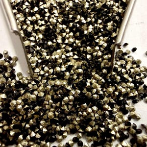 May include: A collection of small, faceted, round rhinestones in alternating metallic gold and solid black colors. The tiny, sparkling gems are poured from a silver-toned metal scoop, creating a dense, textured pile of reflective surfaces.