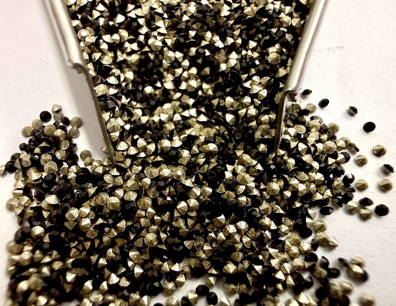 May include: A collection of small, faceted, round rhinestones in alternating metallic gold and solid black colors. The tiny, sparkling gems are poured from a silver-toned metal scoop, creating a dense, textured pile of reflective surfaces.