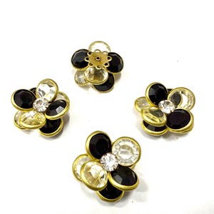 Può includere: Quattro abbellimenti a forma di fiore con base in metallo dorato. Ogni fiore ha pietre nere, trasparenti e bianche. Gli abbellimenti sono disposti su uno sfondo bianco.