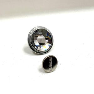 Puede incluir: Un botón grande de cristal transparente facetado con un borde de metal plateado y un botón más pequeño a juego con la palabra "SWAROVSKI" grabada. Los botones están sobre un fondo blanco.