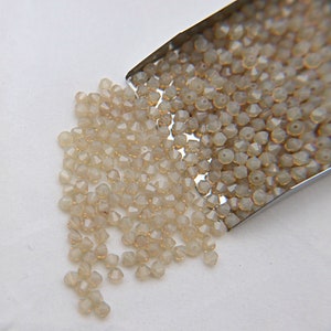 Op de afbeelding: Een close-up van een kleine metalen schep gevuld met heldere, gefacetteerde glazen kralen. De kralen zijn lichtbeige van kleur en hebben een subtiele glans.