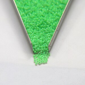 Op de afbeelding: Een metalen bak gevuld met kleine, felgroene glazen kralen. De kralen vallen uit de bak op een wit oppervlak.