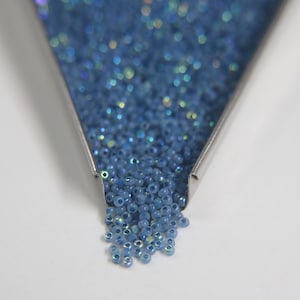 May include: A close-up of a metal funnel filled with small, iridescent blue seed beads. The beads are spilling out of the funnel onto a white surface.