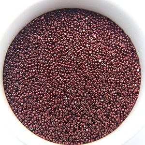 Puede incluir: Una vista de cerca de un tazón lleno de pequeñas cuentas redondas y metálicas. Las cuentas son de color burdeos oscuro con un acabado brillante.