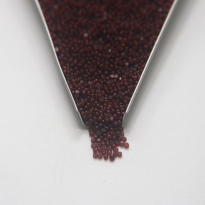 Puede incluir: Primer plano de un pequeño embudo de metal lleno de cuentas de semillas de color rojo oscuro. Las cuentas se derraman del embudo sobre una superficie blanca.