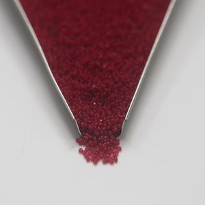 Może przedstawiać: Zbliżenie na metalową łyżkę wypełnioną małymi, czerwonymi koralikami. Koraliki wysypują się z łyżki na białe podłoże.