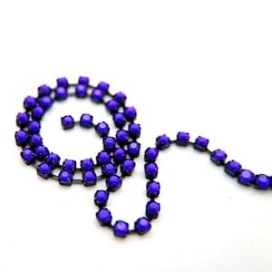 Puede incluir: Primer plano de una cadena de strass púrpura. La cadena está hecha de pequeños strass redondos que están conectados por eslabones de metal negro. La cadena está enrollada en forma de espiral.