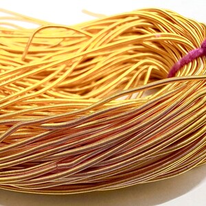 Könnte beinhalten: Ein großes Bündel aus dünnem, flexiblem, goldfarbenem Metalldraht. Der Draht weist eine feine, gewickelte Textur entlang seiner Länge auf und wird durch ein leuchtend pinkfarbenes Garn zusammengehalten, das an einer Seite festgebunden ist.
