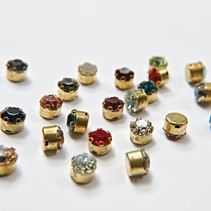 May include: Assortment of colorful rhinestones with gold settings. The rhinestones are in various colors, including red, blue, green, and black.
