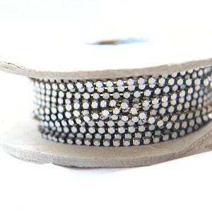 Puede incluir: Un carrete de cadena de pedrer&iacute;a. La cadena est&aacute; hecha de peque&ntilde;as piedras de estr&aacute;s redondas y transparentes engastadas en un metal dorado. La cadena est&aacute; enrollada alrededor de un carrete de cart&oacute;n, creando un elemento decorativo y brillante.