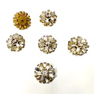 Può includere: Sei bottoni vintage a forma di fiore, con strass e metallo dorato. Ogni bottone presenta uno strass centrale trasparente circondato da strass baguette, creando un motivo a stella. Un bottone mostra il retro con gambo.