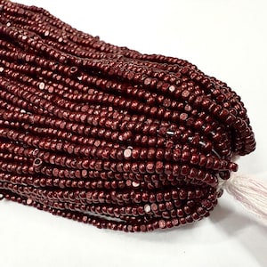 Op de afbeelding: Een bundel kleine, donkerrode kralen, strak aan elkaar geregen. De kralen hebben een licht metallic glans en zijn bevestigd aan een witte draad. De kralen zijn waarschijnlijk voor het maken van sieraden of andere ambachten.