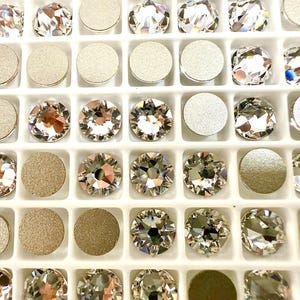 Könnte beinhalten: Nahaufnahme einer weißen Schale gefüllt mit funkelnden, facettierten Kristall-Strasssteinen und metallischen, kreisförmigen Verzierungen. Die Kristalle sind klar mit mehreren Facetten, die das Licht reflektieren. Die Metallteile sind silber- und goldfarben.