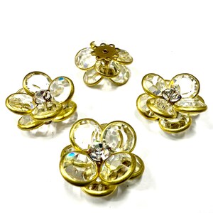 Può includere: Quattro abbellimenti decorativi a forma di fiore. Ogni fiore ha una base color oro con petali trasparenti e sfaccettati e un strass centrale trasparente. Gli abbellimenti sono disposti su uno sfondo bianco.