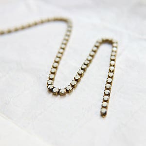 Puede incluir: Un delicado collar con una serie de peque&ntilde;as piedras preciosas blancas y redondas engastadas en una cadena de metal dorado. El collar se muestra sobre una superficie blanca texturizada, creando un contraste. La cadena es flexible y tiene una ligera curva.