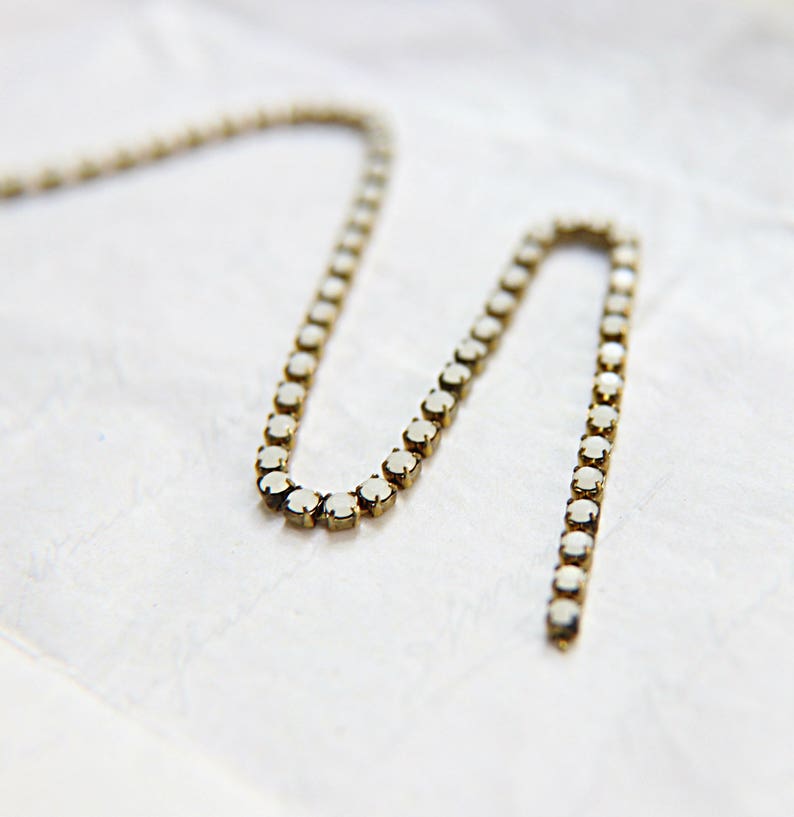 Puede incluir: Un delicado collar con una serie de peque&ntilde;as piedras preciosas blancas y redondas engastadas en una cadena de metal dorado. El collar se muestra sobre una superficie blanca texturizada, creando un contraste. La cadena es flexible y tiene una ligera curva.