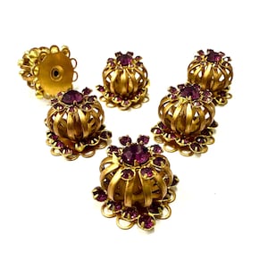 Può includere: Una collezione di sei pomelli per cassetti vintage in metallo dorato con un design floreale. Ogni pomello presenta una gemma centrale viola circondata da pietre viola più piccole e una gabbia metallica traforata decorativa.