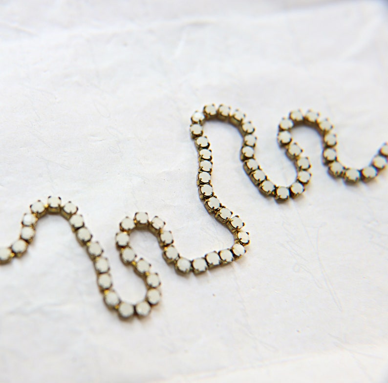 Puede incluir: Primer plano de una cadena de joyer&iacute;a decorativa con peque&ntilde;as piedras preciosas redondas de color blanquecino engastadas en un metal dorado. La cadena est&aacute; dispuesta en un patr&oacute;n ondulado sobre un fondo blanco.