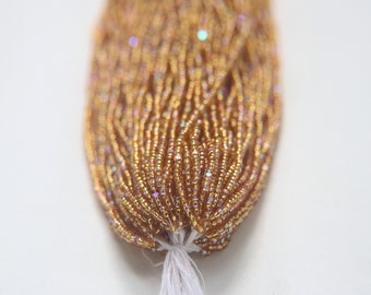 May include: A close-up of a bundle of gold seed beads with iridescent shimmer. The beads are arranged in a tight, compact bundle, with the ends of the strands visible at the bottom.