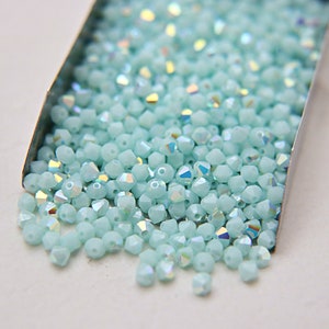 Könnte beinhalten: Nahaufnahme eines Behälters gefüllt mit hellblauen, irisierenden Glasperlen. Die Perlen sind klein und haben eine facettierte Form.