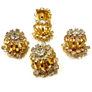 Può includere: Quattro orecchini dorati in stile vintage con un design floreale. Ogni orecchino presenta un cristallo trasparente centrale circondato da strass più piccoli e una struttura a gabbia. Gli orecchini sono su uno sfondo bianco.