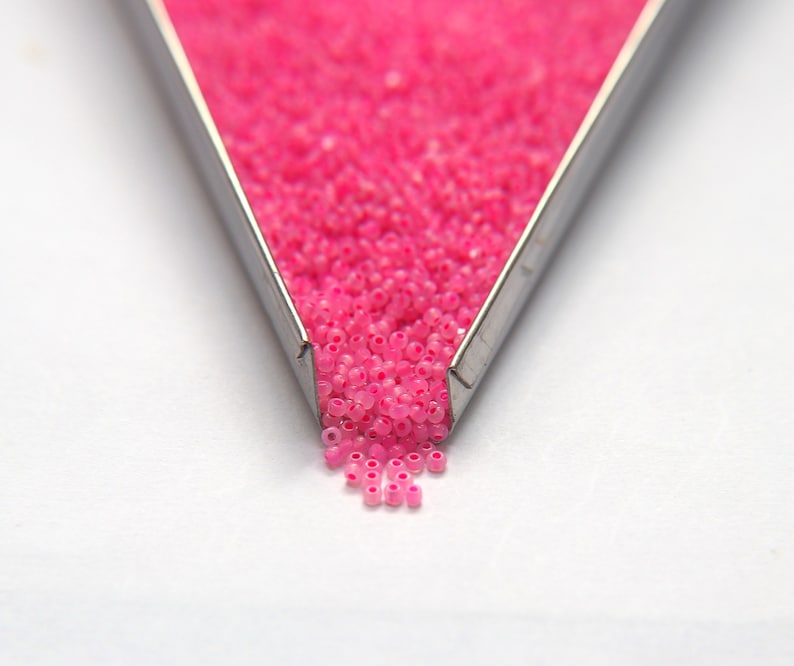 Op de afbeelding: Een metalen bak gevuld met roze zaadkraaltjes die op een wit oppervlak vallen.