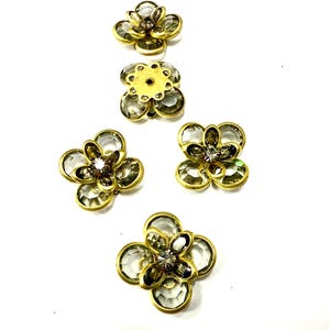 Può includere: Cinque abbellimenti a forma di fiore con petali trasparenti e sfaccettati e un strass centrale. I fiori hanno una base in metallo color oro. Gli abbellimenti sono disposti su uno sfondo bianco.