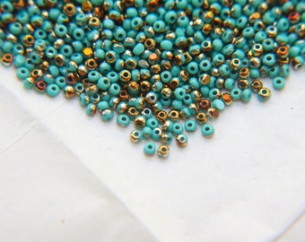 May include: A pile of turquoise and gold seed beads. The beads are small and round, and they are scattered on a white surface.